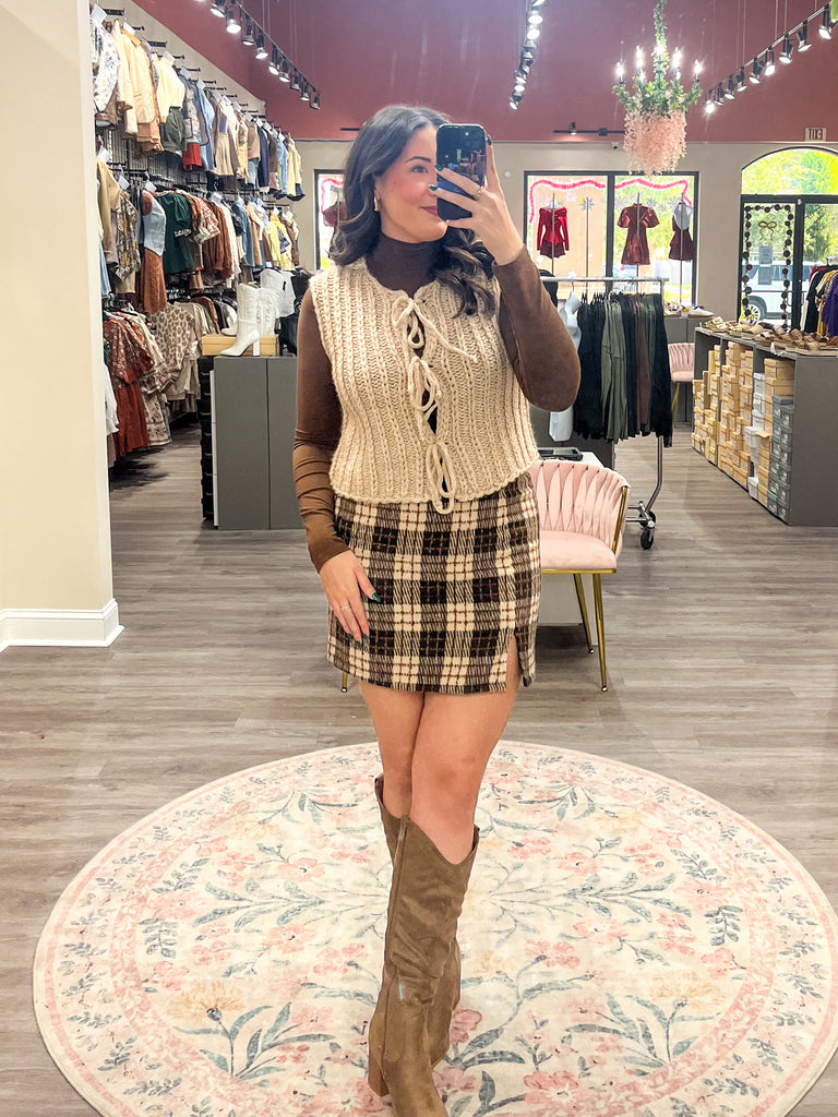Woman taking a mirror selfie in a clothing store wearing a sleeveless knitted top and plaid skirt.