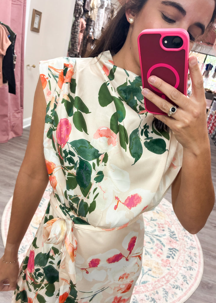 Cream satin maxi dress with bold floral print, high neckline, and waist tie – perfect for weddings or summer events.