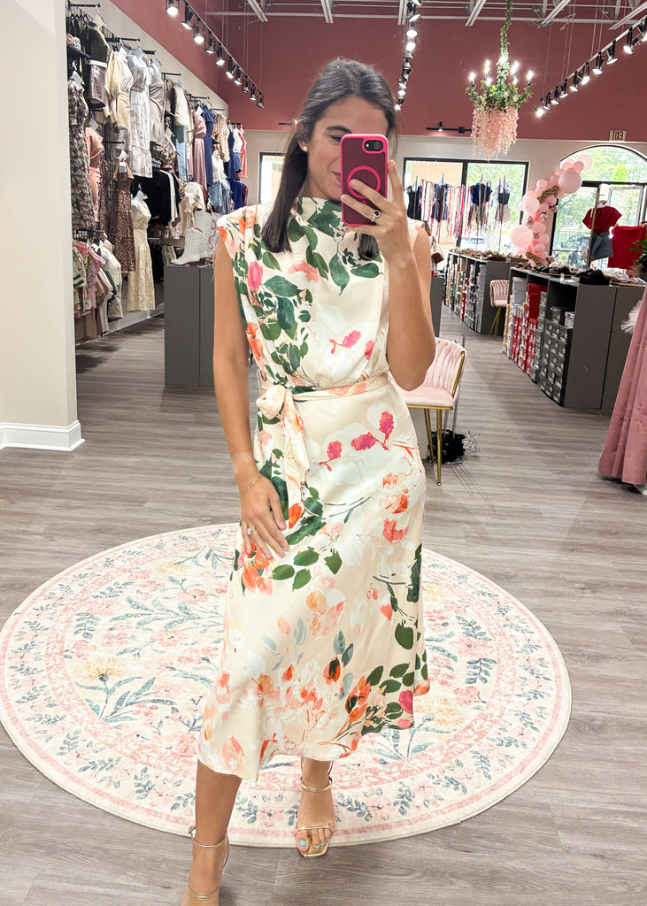Cream satin maxi dress with bold floral print, high neckline, and waist tie – perfect for weddings or summer events.