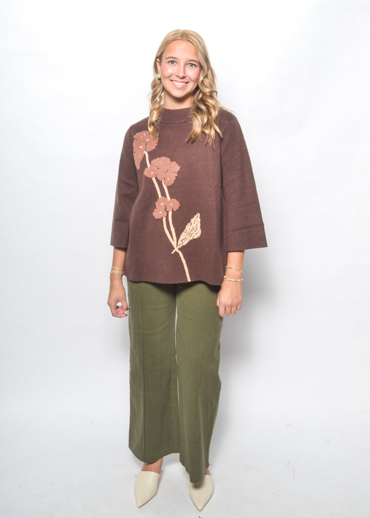 Woman wearing a brown sweater with floral patterns and green pants on a white background