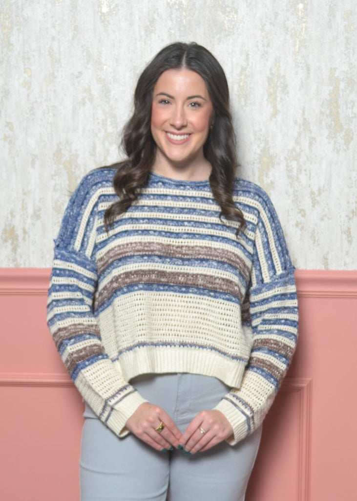 Woman wearing a striped sweater and light blue pants against a pink paneled wall.